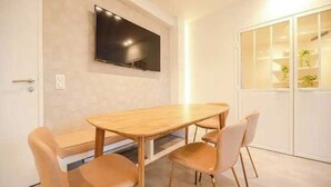 Une pièce moderne avec une table à manger en bois et quatre chaises, un téléviseur à écran plat fixé au mur, et un meuble blanc avec des portes en verre.