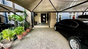 A covered parking area with a black car parked on the right, potted plants on the left, and a building entrance in the background. - Tranquil 1BR w/ Pool Access in Prime Ikeja GRA (Ikeja)
