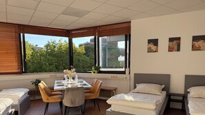 A modern hotel room with a dining table set for two, a large window with a view of greenery, a bed with a grey headboard, and a small bedside table. - City Center Retreat/ 1 min Train Station (Köln)