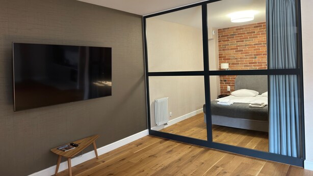 A modern hotel room with a large flat-screen TV, a small wooden side table, a sliding glass door leading to a bedroom with a brick wall, and a ceiling light fixture. - Browar Hevelius Comfort Apartments by GA (Gdańsk)