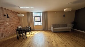 A spacious living room with a wooden floor, a sofa, a dining table with chairs, and a large window with curtains. - Browar Hevelius Comfort Apartments by GA (Gdańsk)