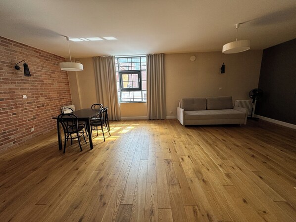 Ein geräumiges Wohnzimmer mit Holzboden, einer Couch, einem Esstisch mit Stühlen und einem großen Fenster mit Vorhängen.