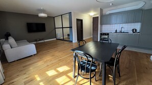 A modern living room with a sofa, a dining table with chairs, a kitchen area with cabinets and appliances, and a large window allowing natural light. - Browar Hevelius Comfort Apartments by GA (Gdańsk)