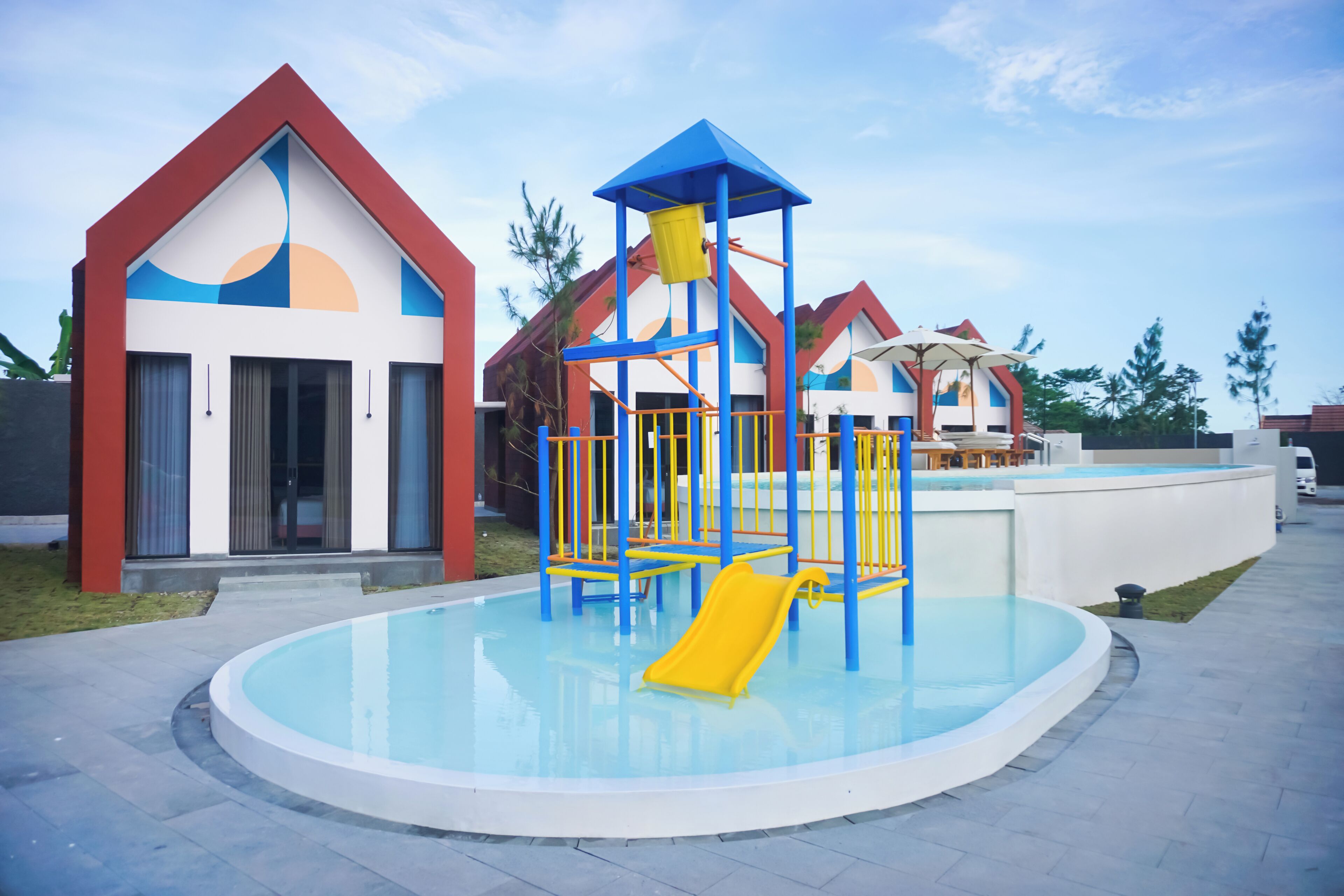 A playground with a slide and climbing structures, surrounded by a pool and modern buildings with unique architectural designs.