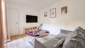 Un salon moderne avec une table de salle à manger ronde, préparée pour quatre personnes, un canapé gris, un téléviseur à écran plat fixé au mur et une porte donnant sur une autre pièce.