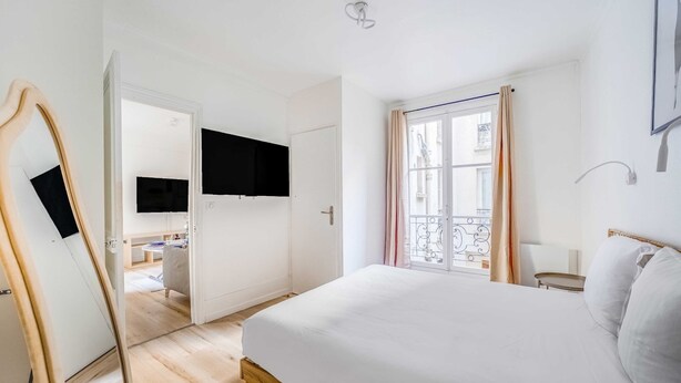 Une chambre à coucher avec un lit, un téléviseur fixé au mur, une fenêtre avec des rideaux et une porte donnant sur une autre pièce.
