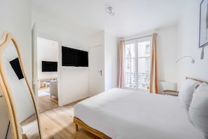 Un dormitorio con cama, televisión montada en la pared, ventana con cortinas y puerta que da a otra habitación.