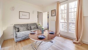Un salon moderne avec une table ronde en verre, disposée pour accueillir quatre personnes, un canapé gris et une grande fenêtre agrémentée de rideaux.