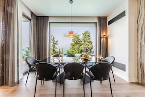 Un coin repas avec une table en verre, quatre chaises, un luminaire suspendu et une vue sur des arbres et une étendue d’eau, le tout visible à travers de grandes fenêtres.