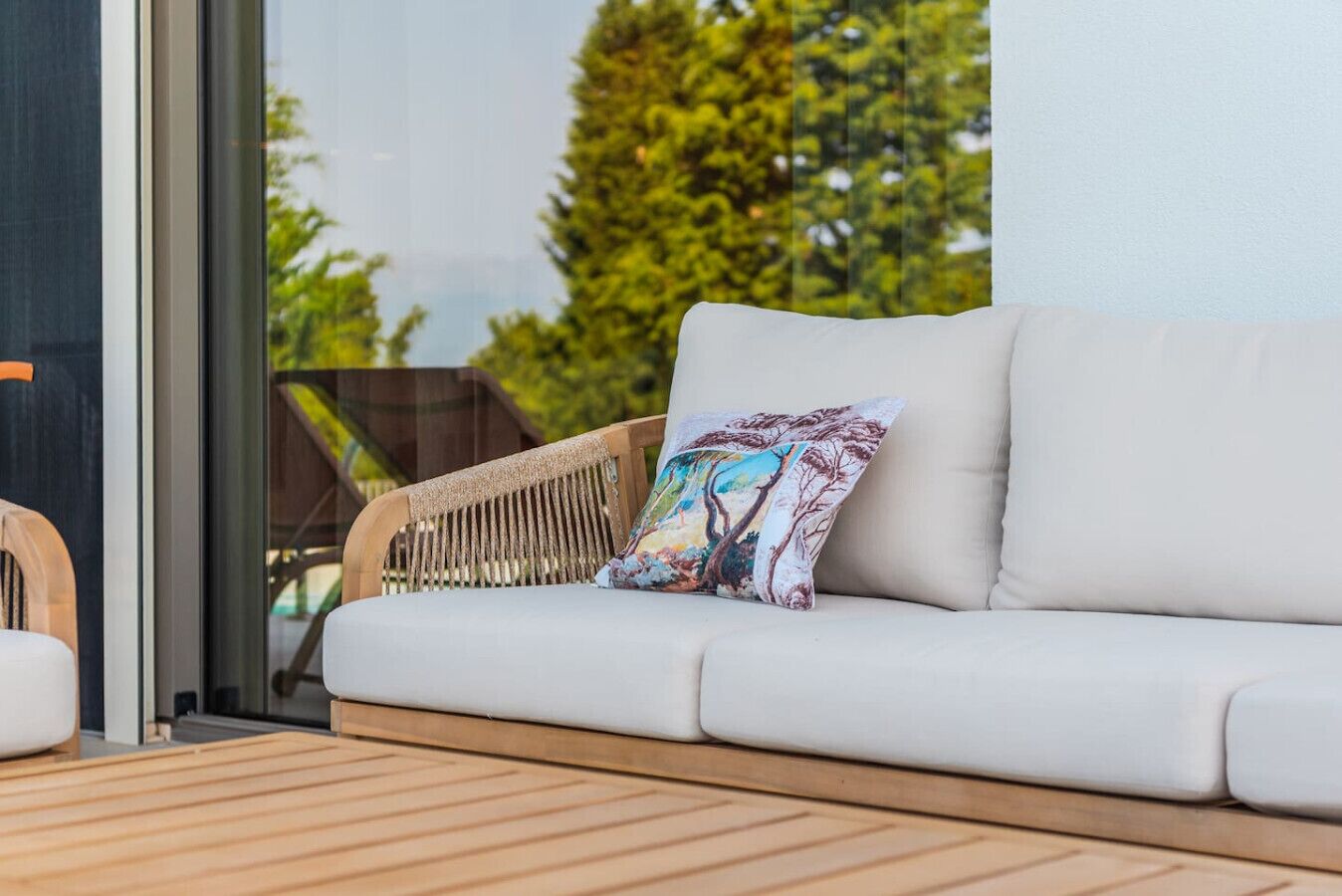Un sofá blanco para exterior con un cojín colorido, una silla de mimbre y una terraza de madera.