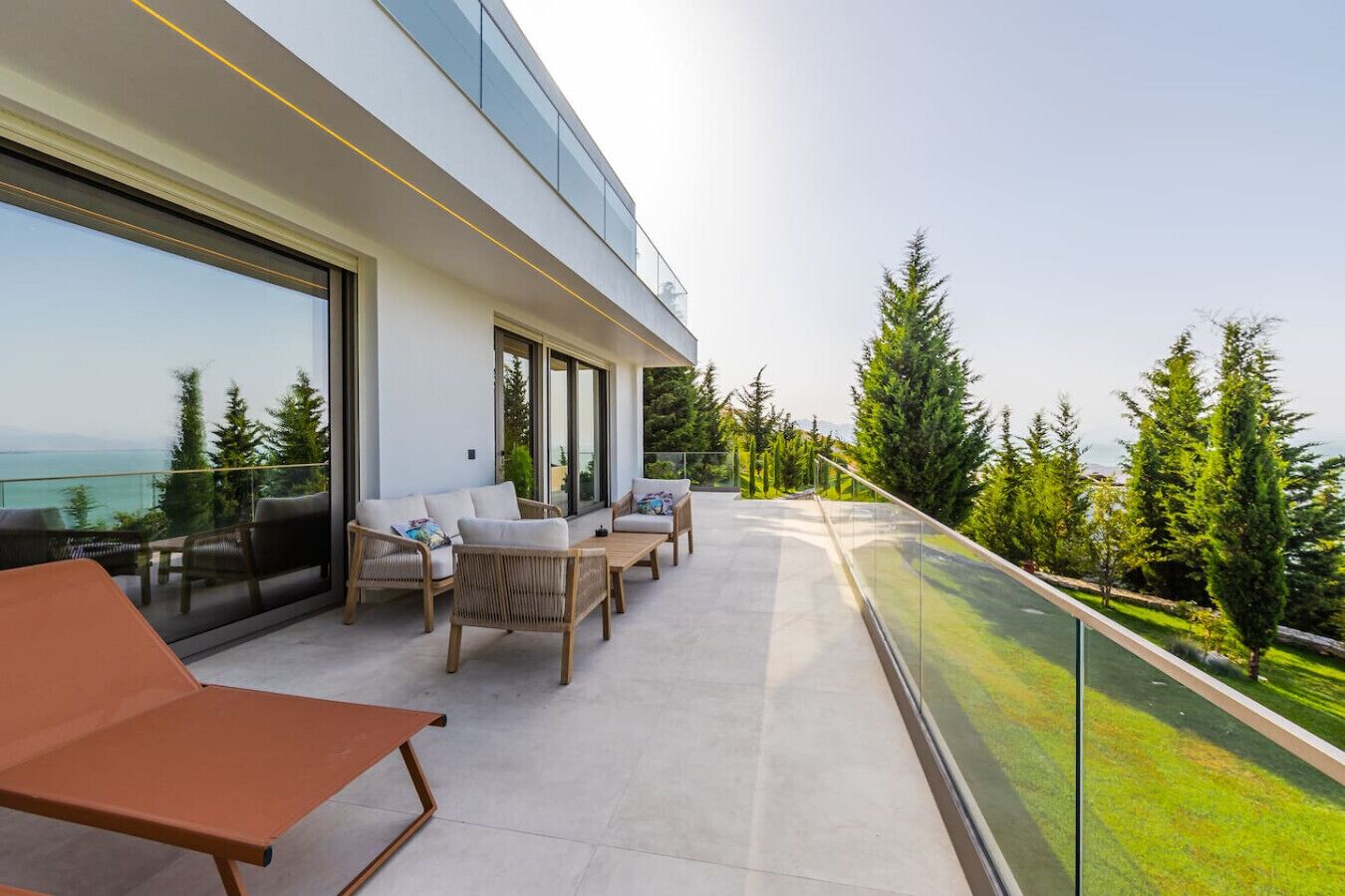 A modern outdoor balcony with a seating area, large windows, and a view of the sea.