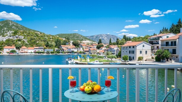 A balcony with a table set with fruit and drinks, overlooking a lake with boats and buildings in the background.