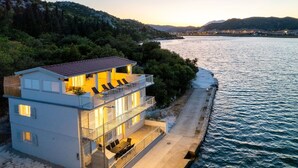 A modern two-story house with a balcony overlooking a body of water, surrounded by greenery.