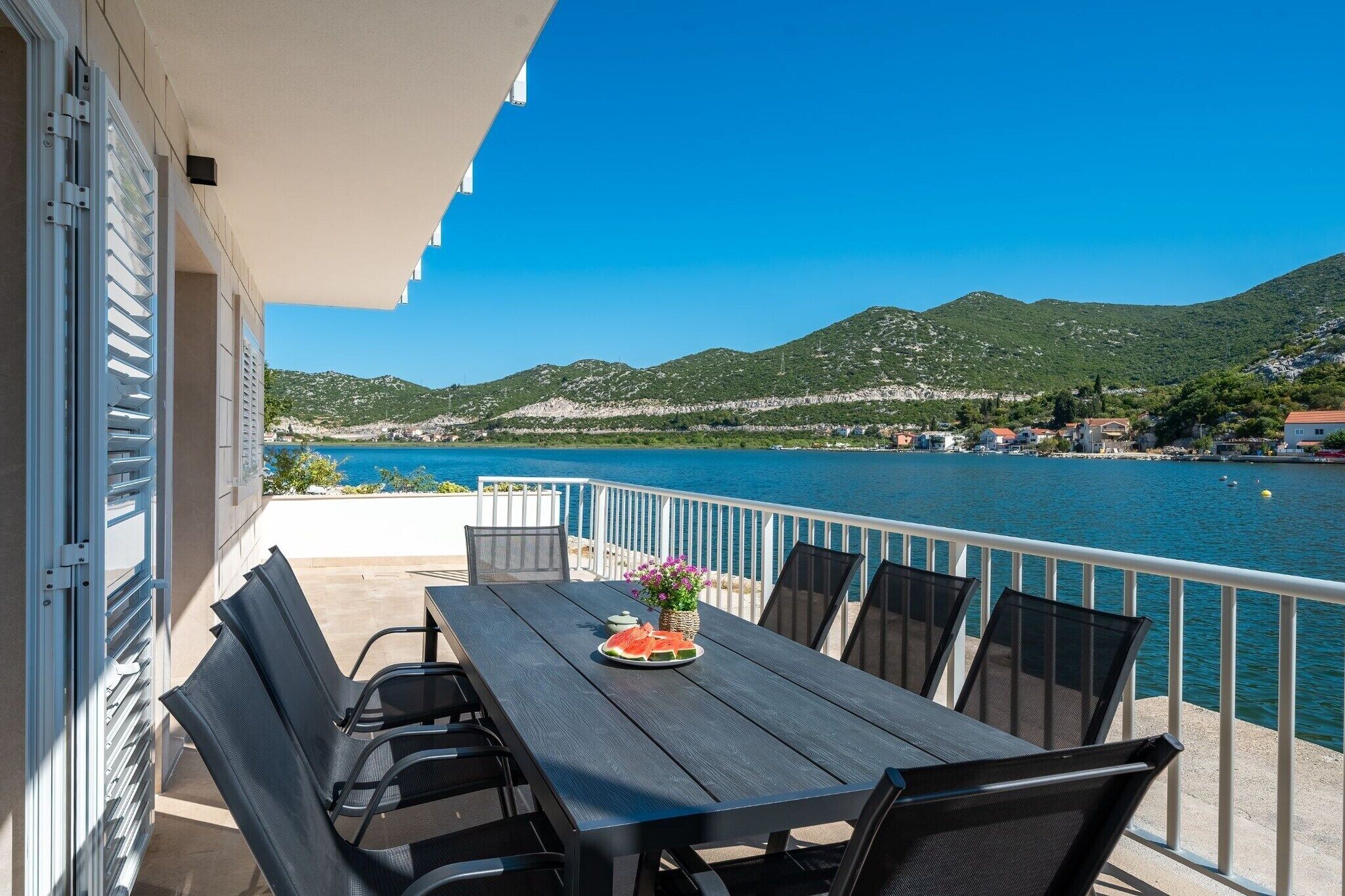 Un balcon avec une table dressée pour deux personnes, offrant une vue sur une étendue d’eau et un paysage montagneux.