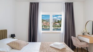 A hotel room with a bed, a desk, a chair, a lamp, a mirror, and a window with a view of a town and mountains.