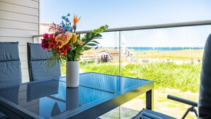 A balcony with a glass table, a potted plant with flowers, and a view of the beach and houses.