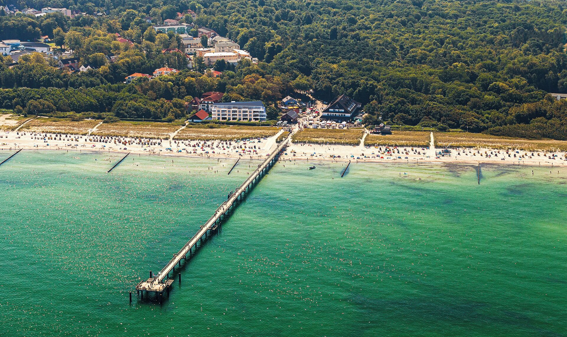 Eine Luftaufnahme einer Küstenlandschaft mit einem Pier, der ins Wasser ragt, einem Strand voller Menschen und Gebäuden im Hintergrund.