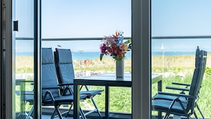 A room with a view of the beach, a table with a vase of flowers, and two chairs.