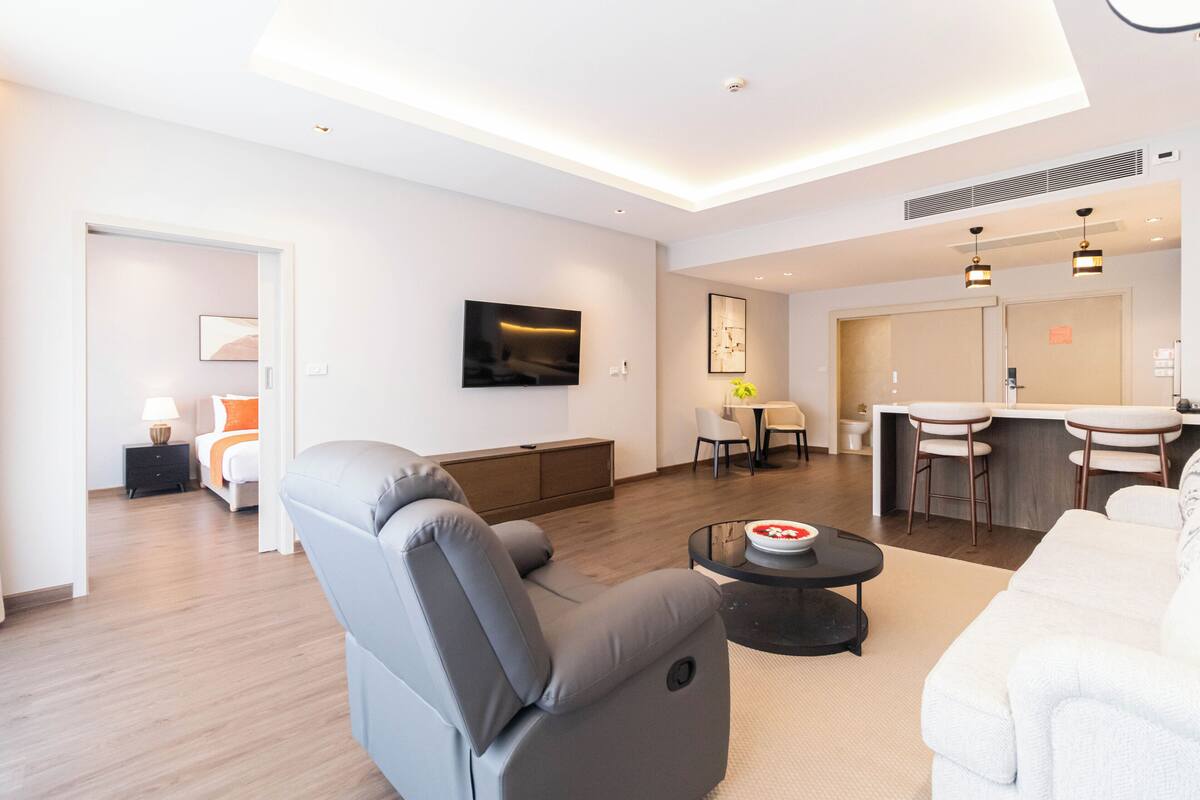A modern living room with a grey sofa, a white couch, a dining area with a table and chairs, and a kitchen area with a bar.