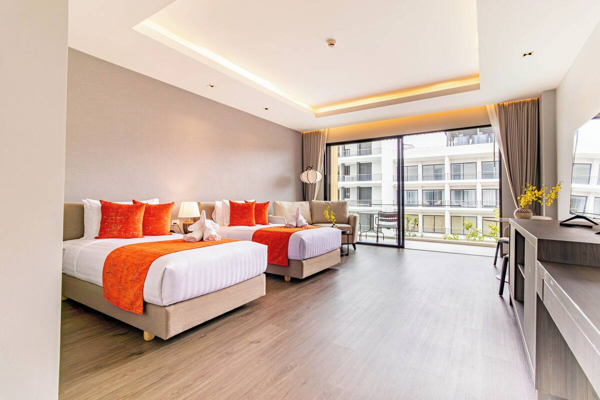 A hotel room with two beds, a desk, a chair, a lamp, and a view of a building.