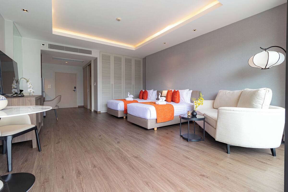 A modern hotel room with two beds, a sofa, a dining table, and a small side table.
