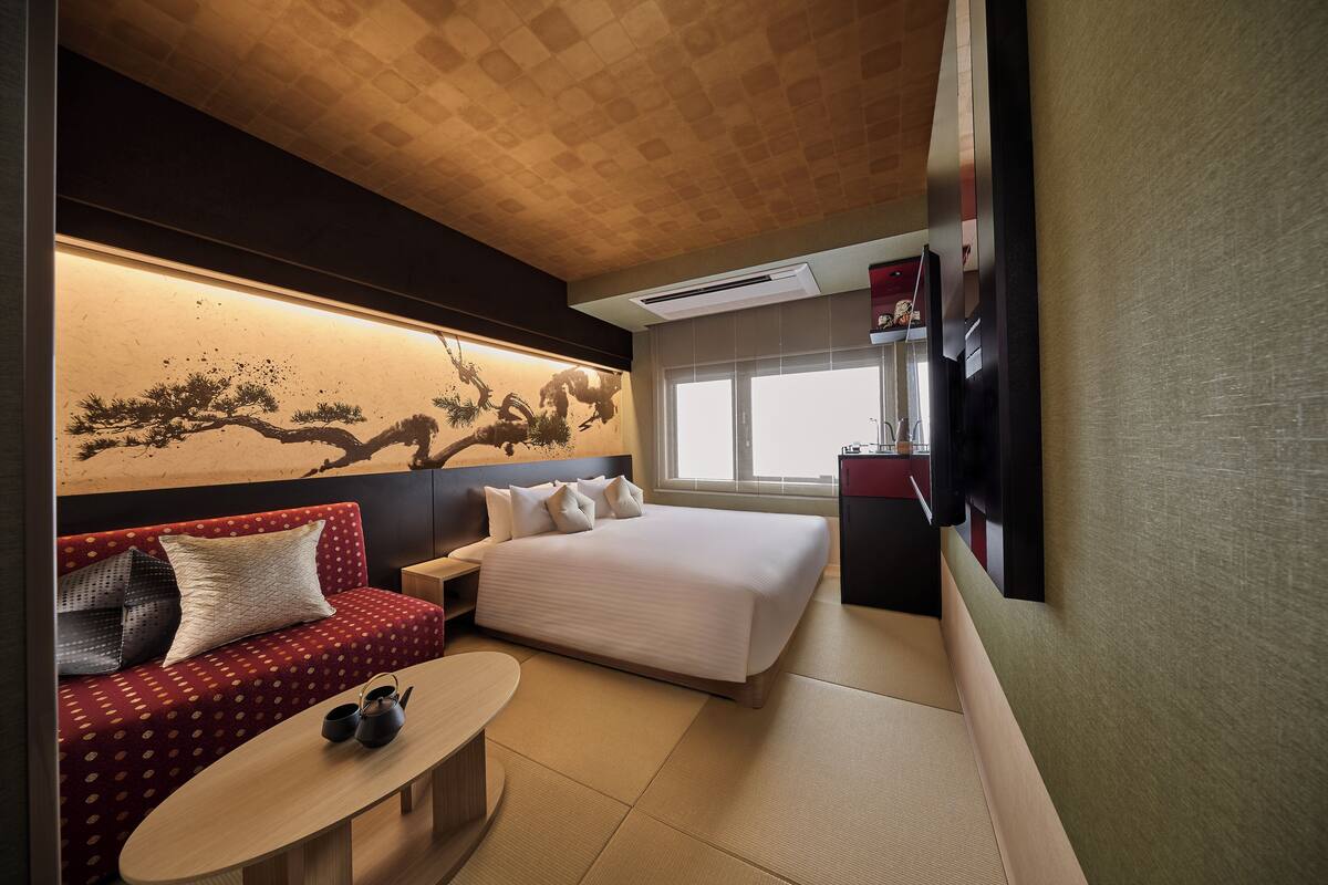 A hotel room with a bed, a sofa, a small table, and a wall-mounted TV.