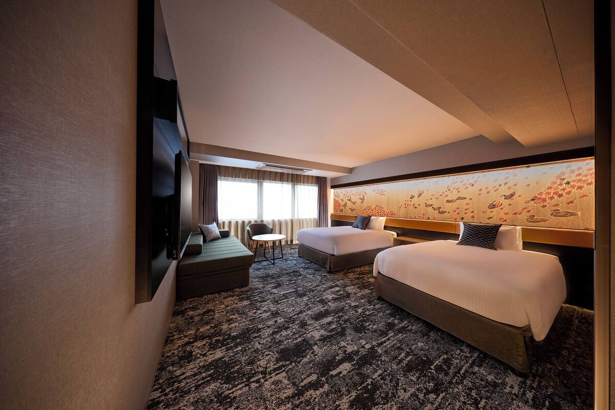 A hotel room with two beds, a sofa, a small table, and a wall-mounted TV.