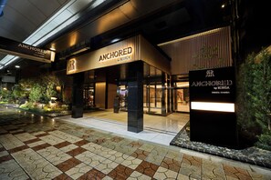 Entrada de un hotel con piso de baldosas, un letrero que dice 'ANCHORED by RIHGA' y fachada de edificio moderno.