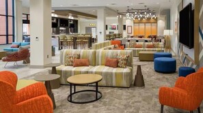 Een moderne hotellobby met een zithoek met banken en poefs, een bar en een groot televisiescherm.