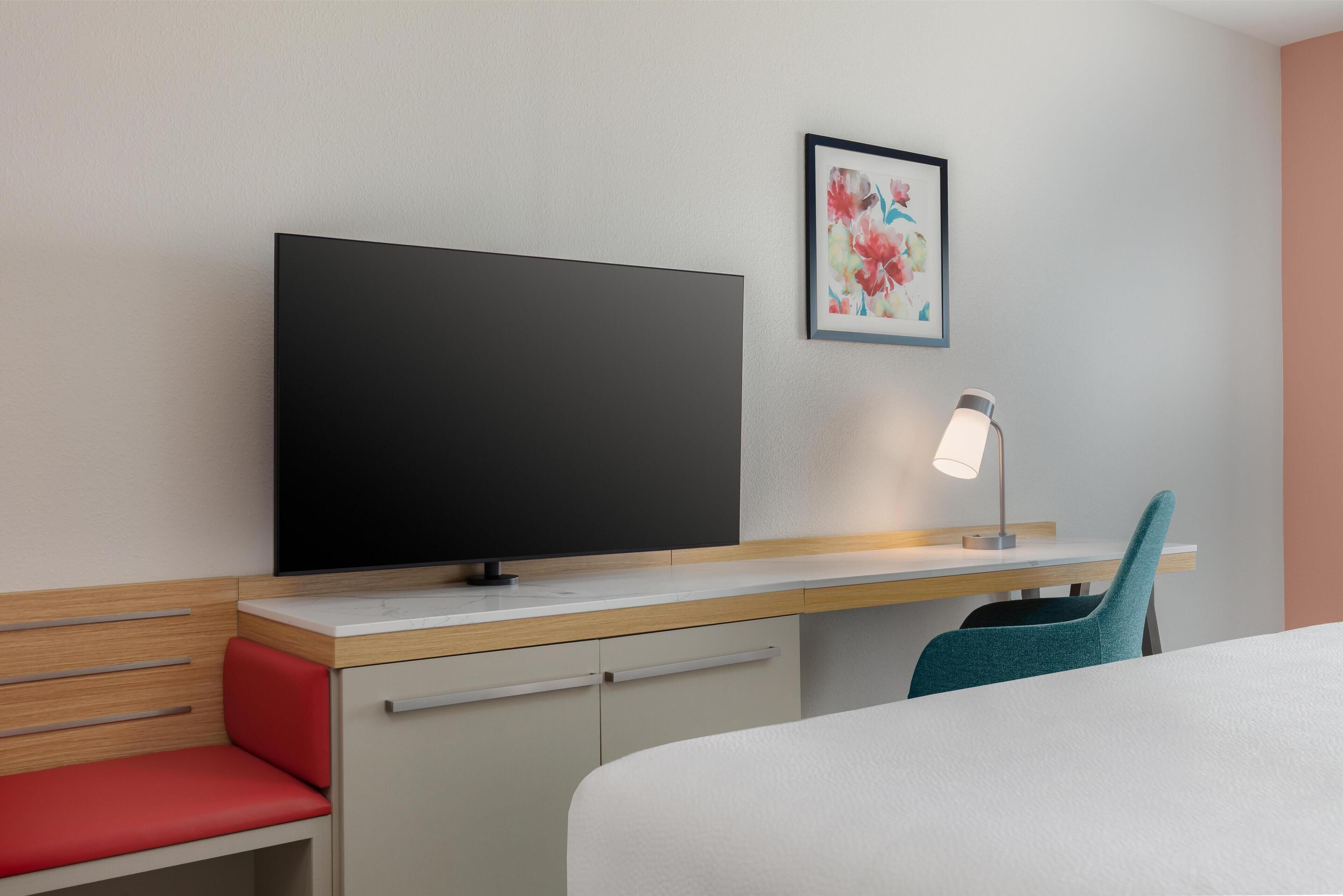 Una habitación de hotel con cama, escritorio con televisor, silla, lámpara de escritorio y un cuadro en la pared.