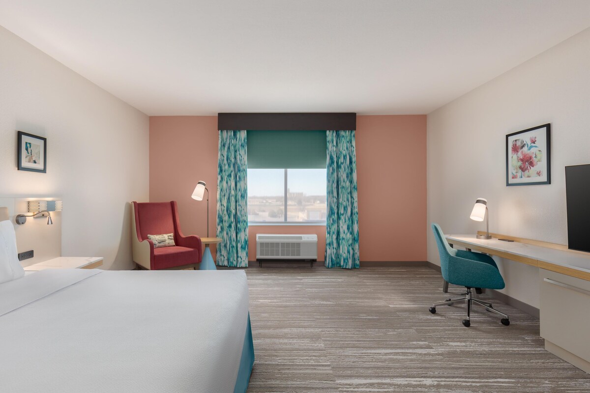 Habitación de hotel con cama, escritorio, silla, equipo de aire acondicionado y ventana con cortinas.