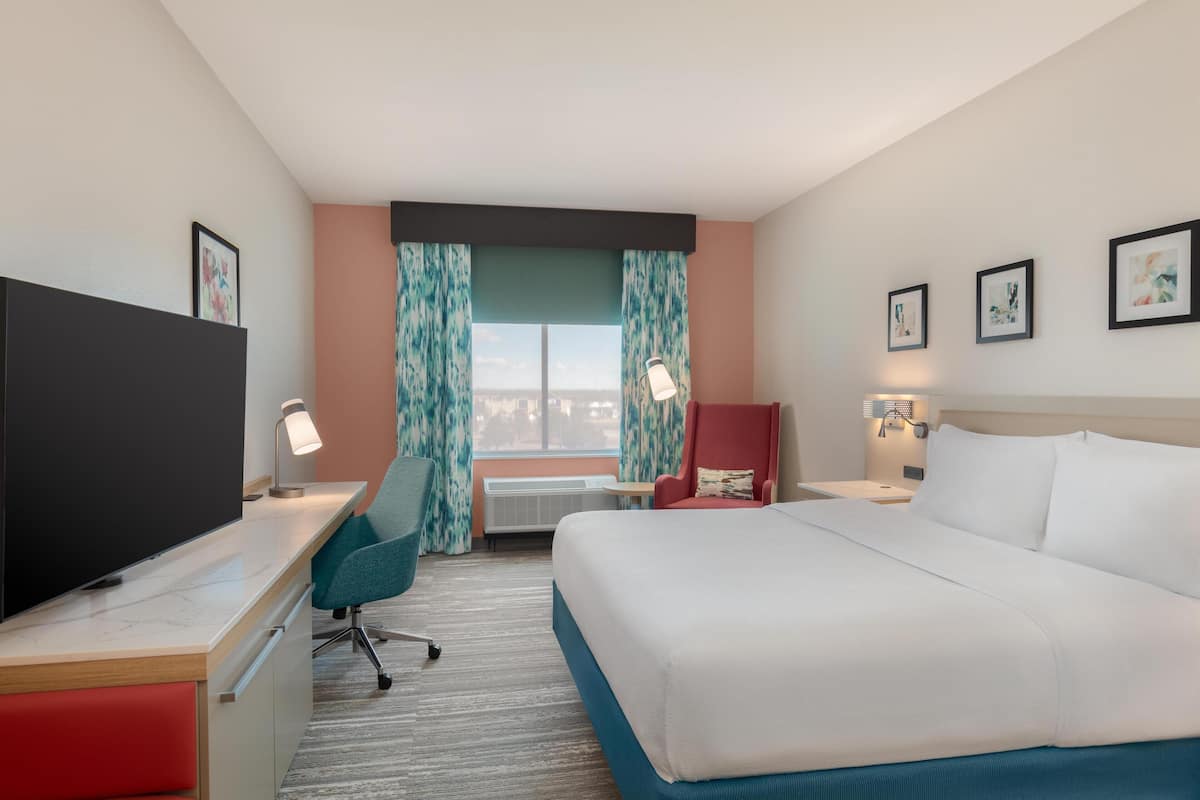 A hotel room with a bed, desk, TV, chair, and a window with curtains.