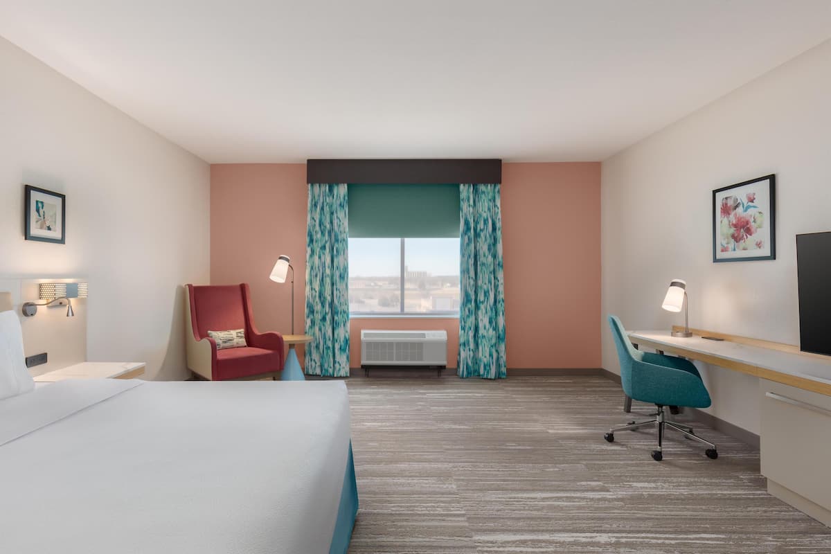 A hotel room with a bed, desk, chair, air conditioning unit, and a window with curtains.