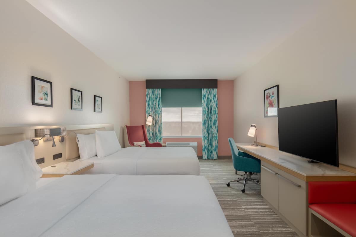 A hotel room with two beds, a desk, a TV, and a window with curtains.