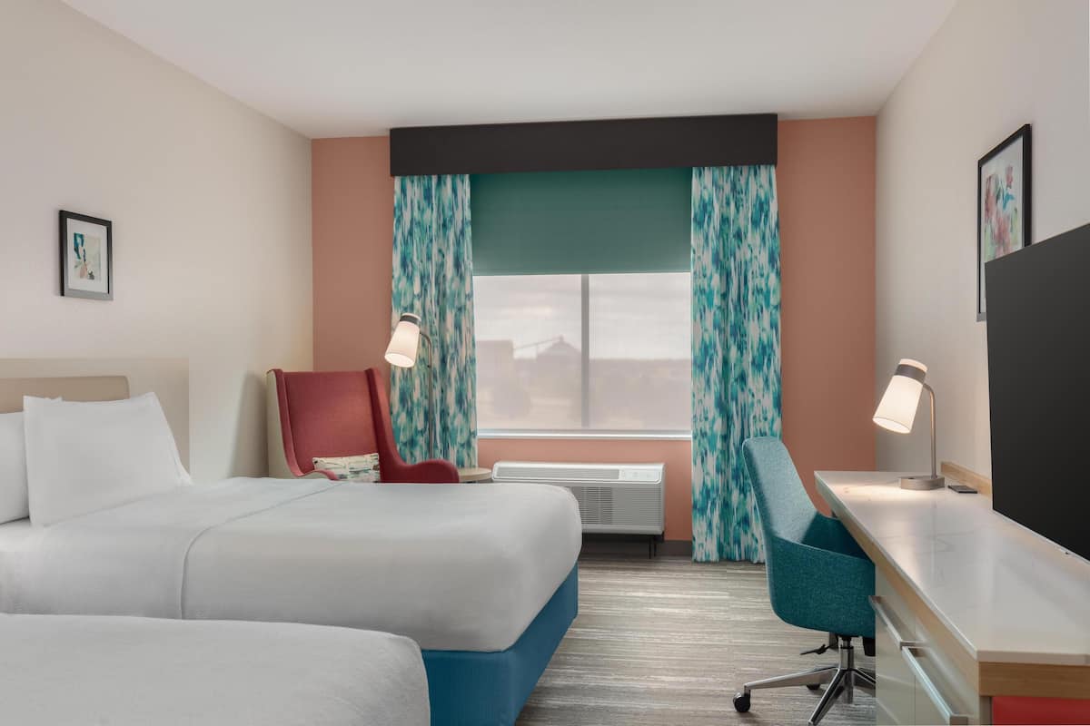 A hotel room with a bed, a desk, a chair, a window with curtains, and an air conditioning unit.
