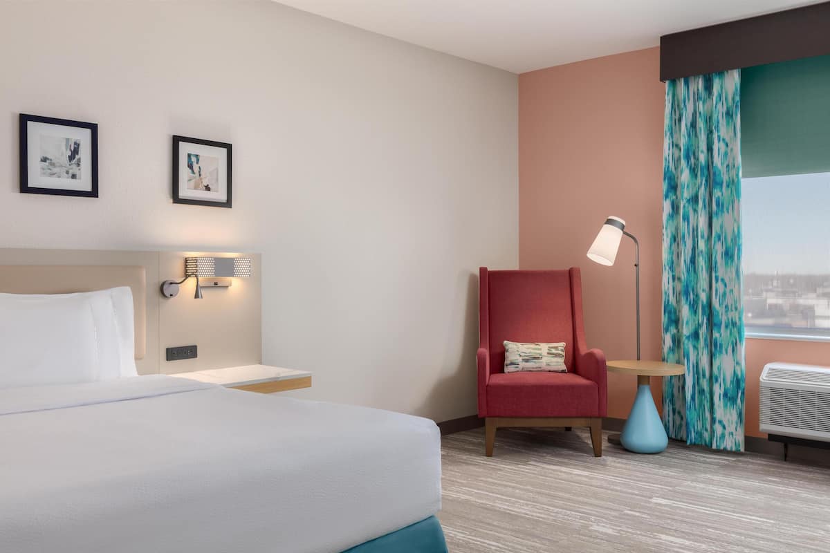 A hotel room with a bed, a red armchair, a small table, a lamp, and a window with curtains.