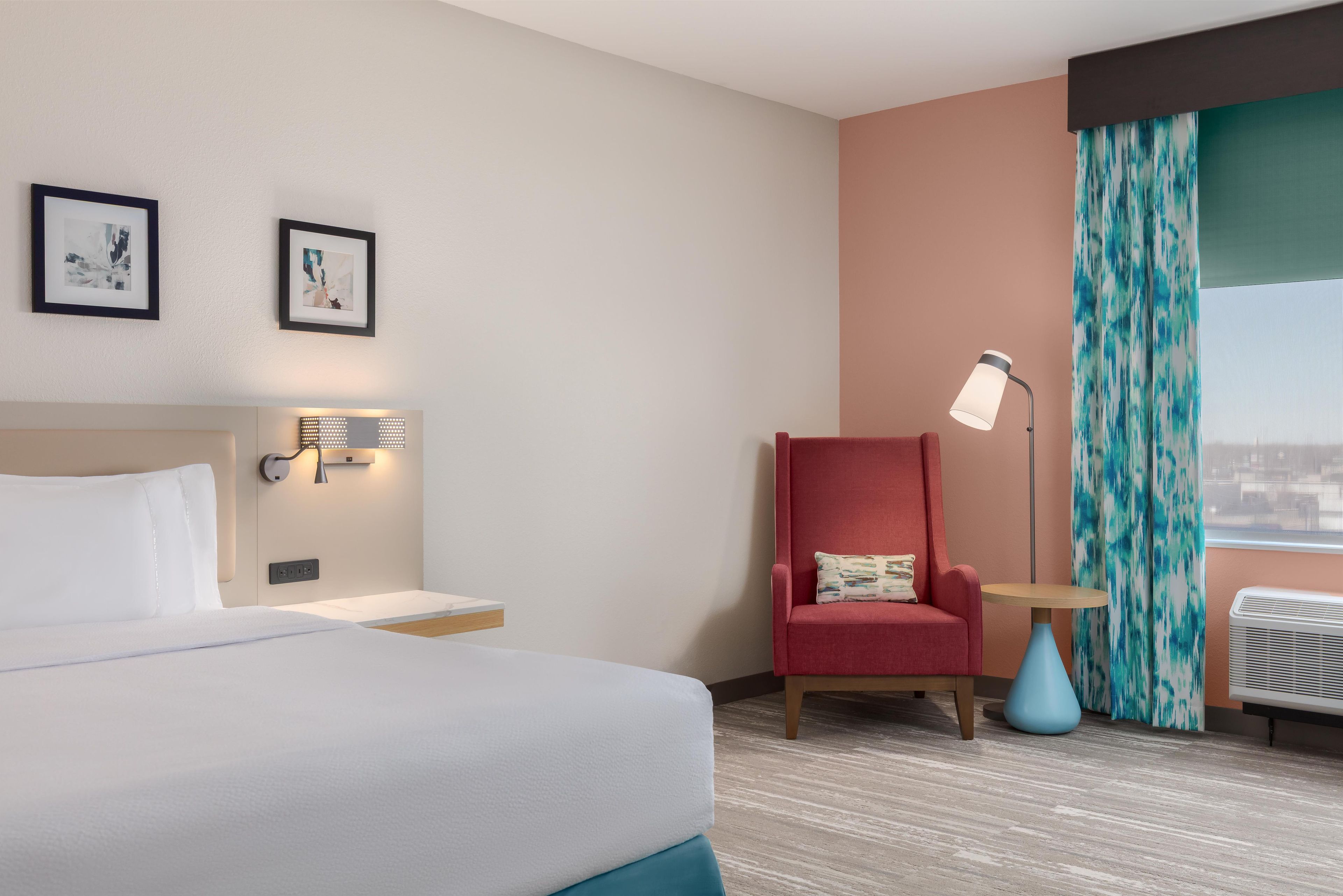 A hotel room with a bed, a red armchair, a small table, a lamp, and a window with curtains.