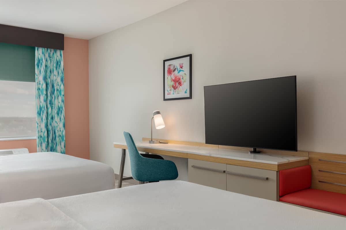 A hotel room with two beds, a desk with a TV, a lamp, and a window with curtains.