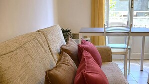 Un sofá color beige con tres cojines, una mesita y una silla en una habitación con ventana y vista a un edificio.