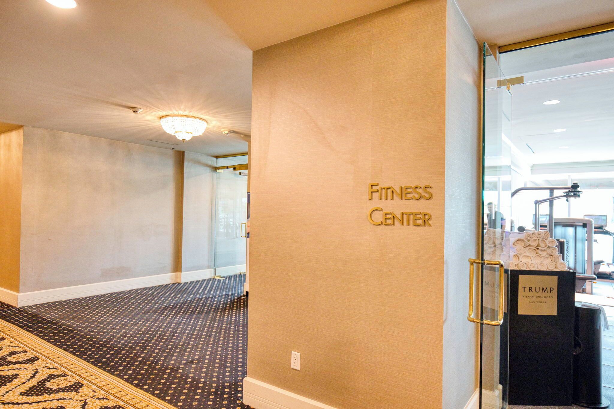 A hotel lobby with a fitness center sign on the wall, a Trump-branded trash bin, and a patterned carpet.