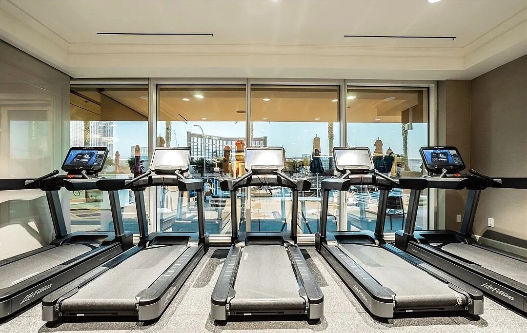 Uma academia com esteiras e equipamentos de exercícios, grandes janelas com vista para prédios e uma piscina.