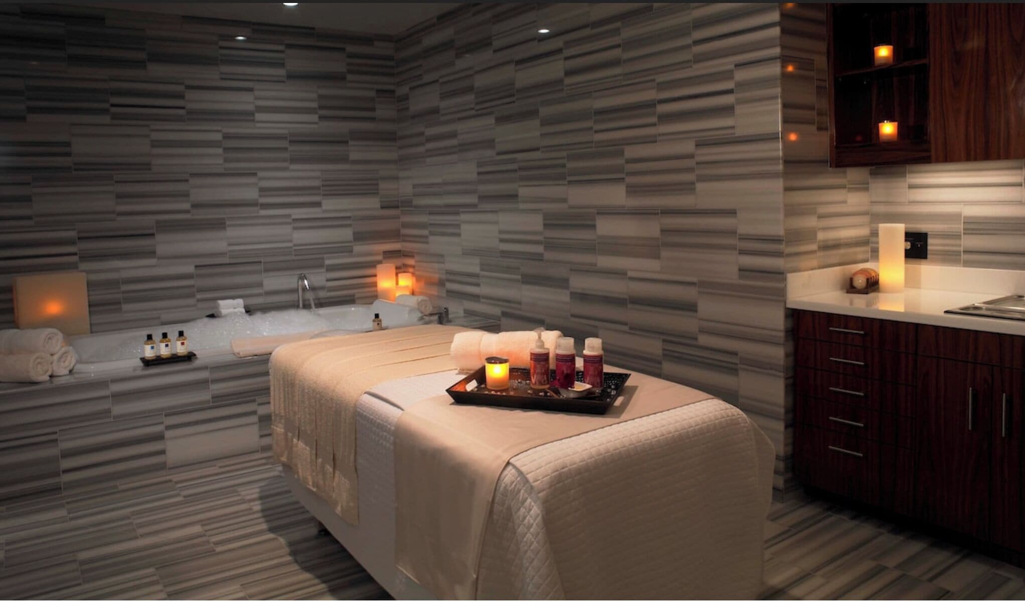 A modern spa room with a large bathtub, a massage table, and a tray of candles and bottles.