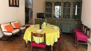 A dining area with a round table covered in yellow, surrounded by wooden chairs with red cushions. A green cabinet with glass doors is in the background.