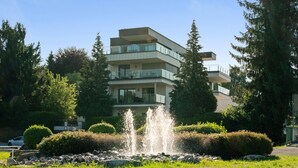 Un edificio residencial de varios pisos con balcones, rodeado de exuberante vegetación y una fuente.