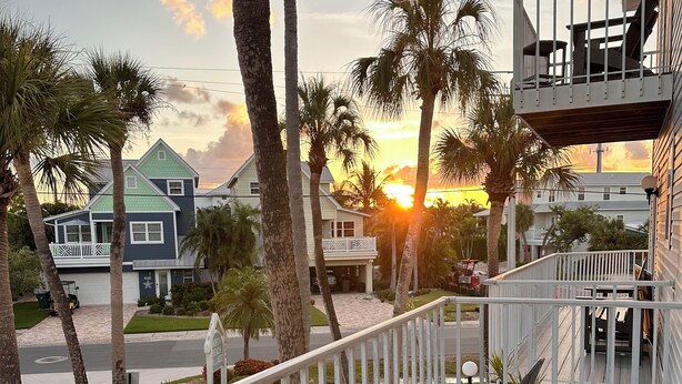 A wooden deck with chairs, overlooking a street with houses and palm trees during sunset.