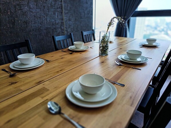 A long wooden dining table set with white bowls and plates, a glass vase with dried flowers, and a view of the city through a window.