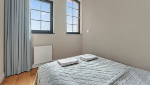 A bedroom with a bed, two windows, a radiator, and a curtain. - Browar Hevelius Apartments by GA (Gdańsk)