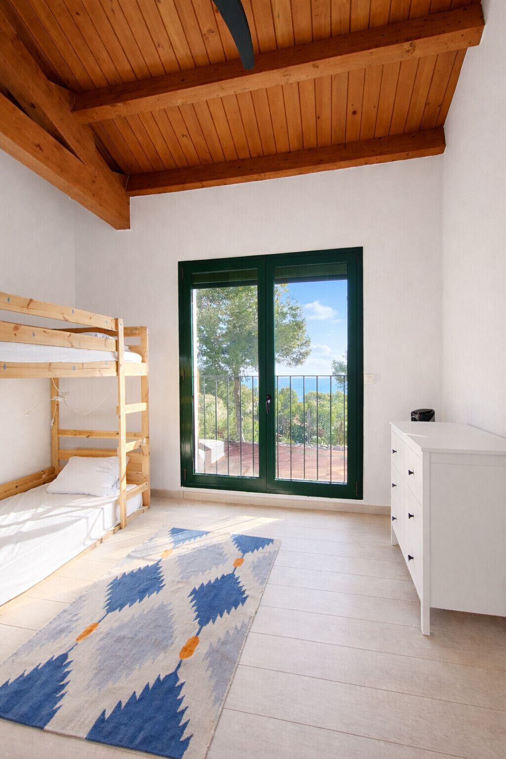 A room with a bunk bed, a white dresser, a patterned rug, and a large sliding glass door with a view of trees and a fence.