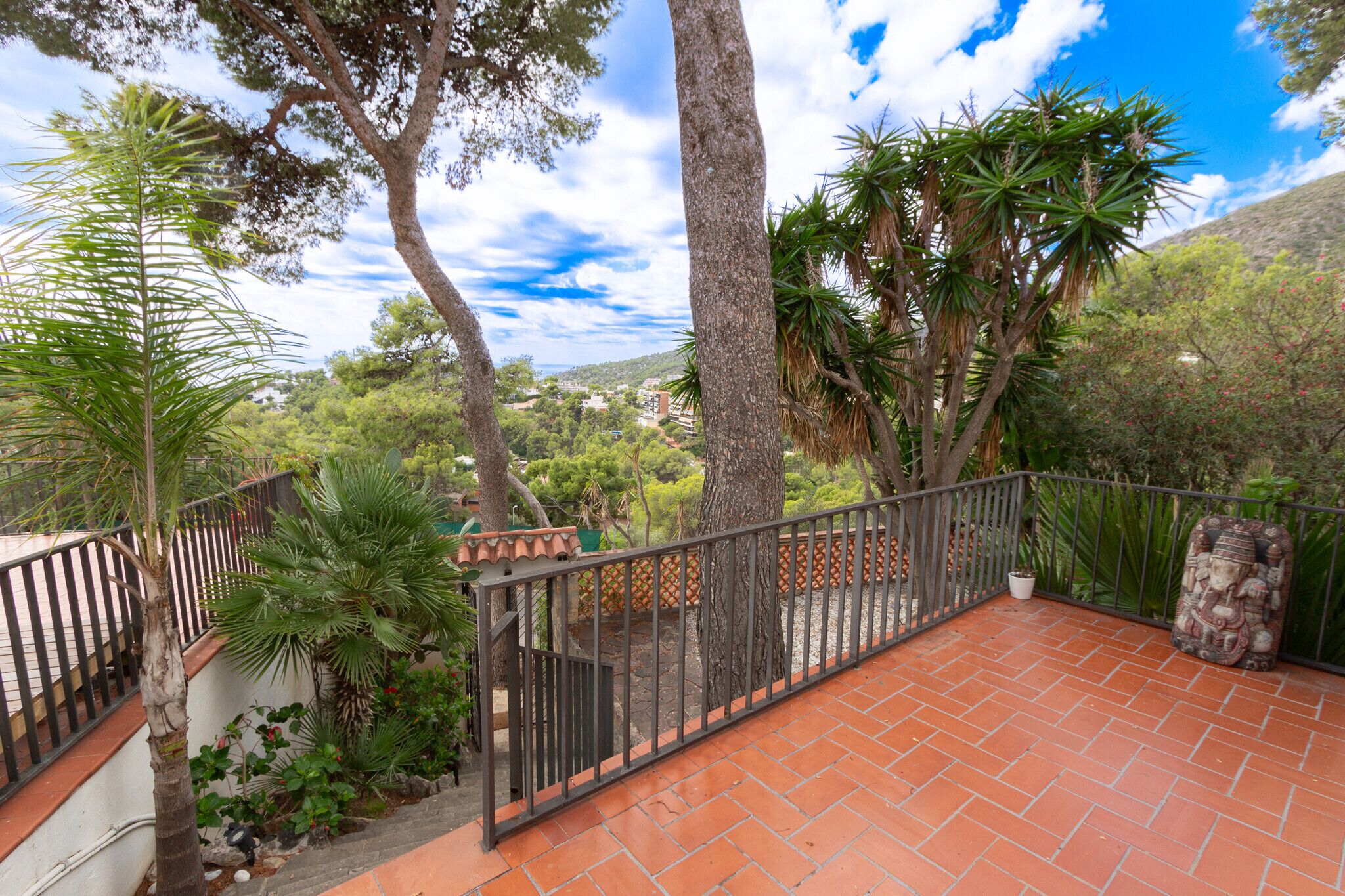 A terracotta-tiled patio with a metal railing, surrounded by various trees and plants.