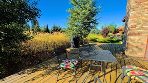 Une terrasse en bois, avec des chaises et une table en métal, entourée de verdure et surplombant un ciel bleu azur.